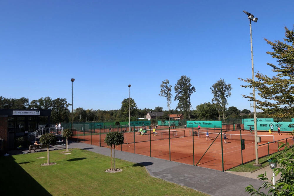 Blick auf das Vereinsheim und die angrenzende Tennisanlage des VfL Grafenwald unter klarem LED Flutlicht.